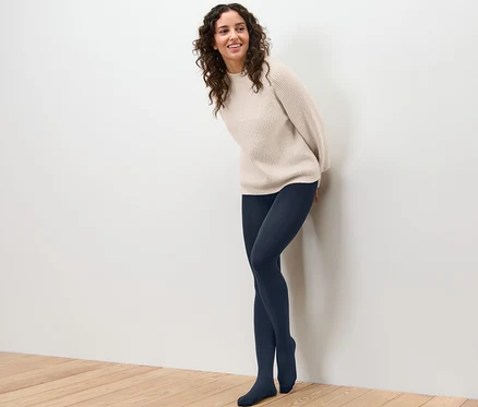 Lächelnde Frau, die sich an eine Wand lehnt, trägt eine Thermostrumpfhose und einen Pullover.