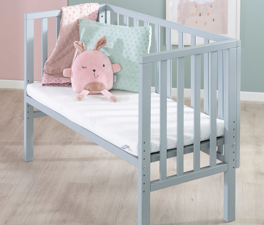 Graues Kinderbett mit rosafarbenem Plüschhasen, rosa Decke und grünem Kissen.