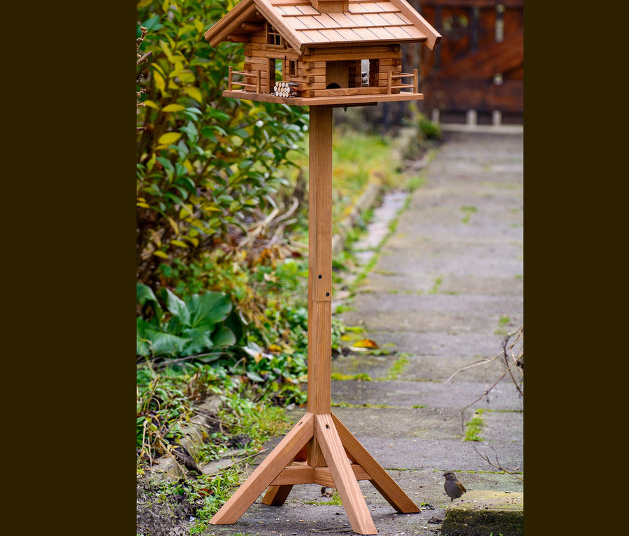 Hölzernes Vogelfutterhaus »Tirol« steht in einem Garten neben einem Busch und ein Vogel steht am Boden daneben.