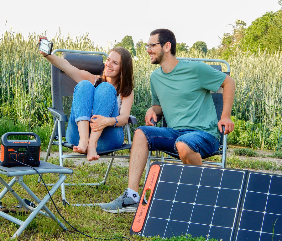 Eine Frau und ein Mann sitzen draußen auf Stühlen. Die Frau hält ein Telefon und macht ein Selfie. Auf einem Tisch stehen ein Jackery Explorer 240 und ein Solarpanel Jackery SolarSaga 100.