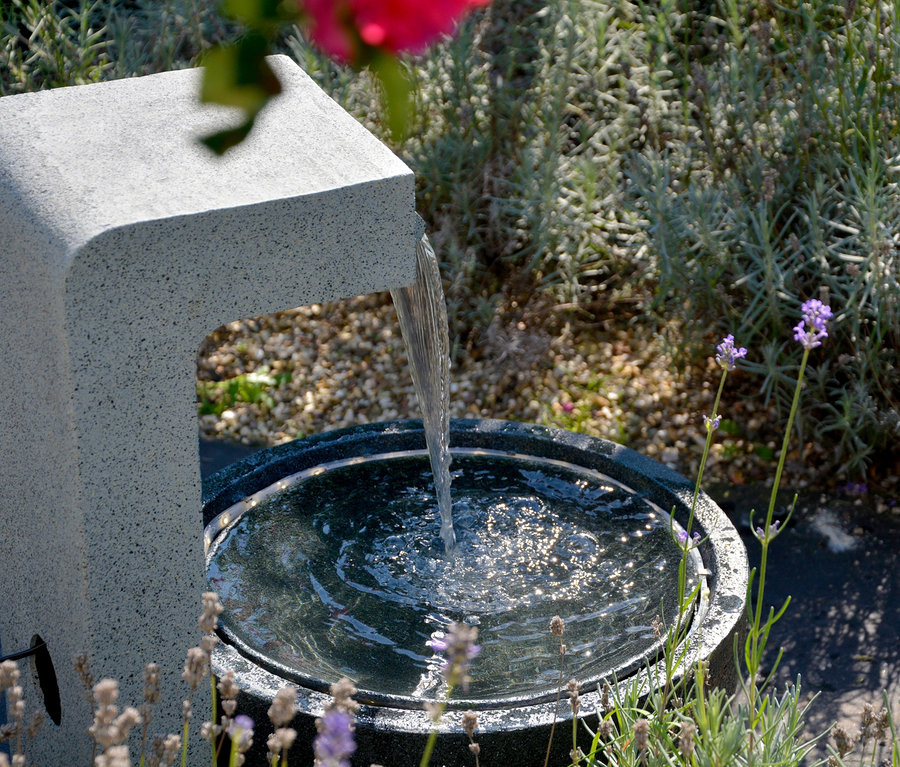 Wasser fließt aus dem steinernen Dobar Gartenbrunnen mit Schale in eine runde Schale inmitten eines Gartens.