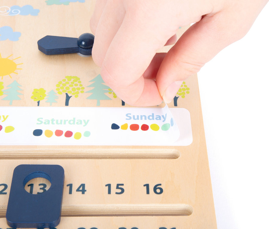 Eine Hand löst einen Aufkleber mit der Aufschrift Sonntag von einem hölzernen Lehrmittel small foot Lernuhr mit Kalender »Educate«.