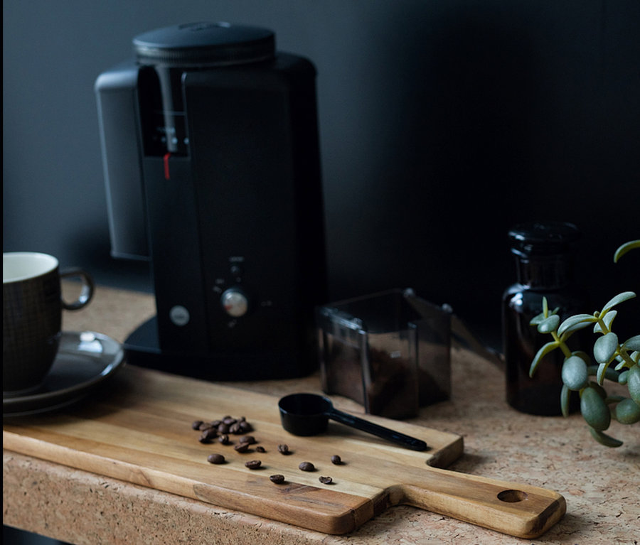 Kaffeebohnen liegen auf einem Holzbrett neben einem schwarzen Messlöffel und der Wilfa Kaffeemühle »Svart Aroma«.