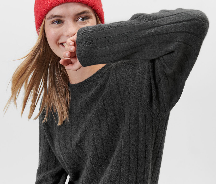 Eine lächelnde Frau trägt ein graues, geripptes Strickkleid und eine rote Mütze.