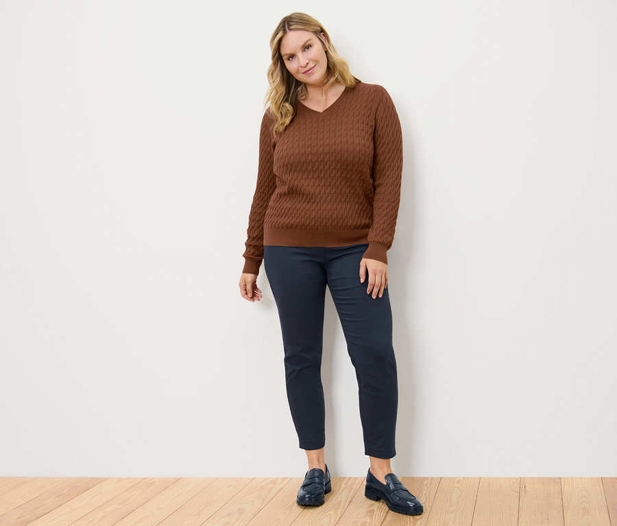 Frau posiert in einem cognacfarbenen Zopfstrickpullover und einer dunkelblauen Stretchhose.