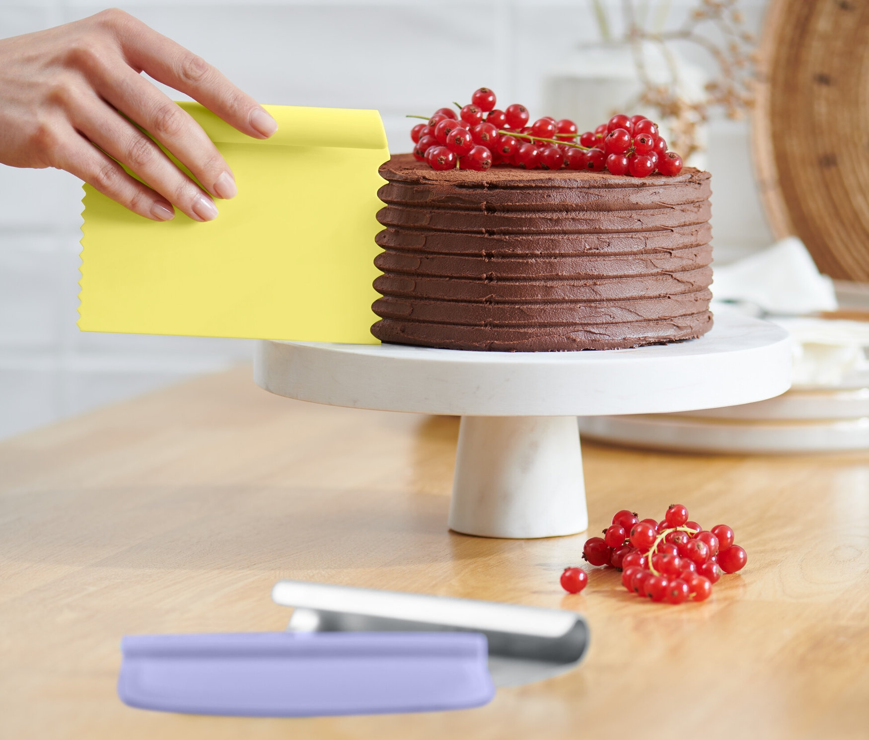 Eine Hand benutzt einen gelben Schaber aus einem Backhelfer-Set, um einen Schokoladenkuchen mit roten Johannisbeeren auf einem weißen Sockel zu bearbeiten.