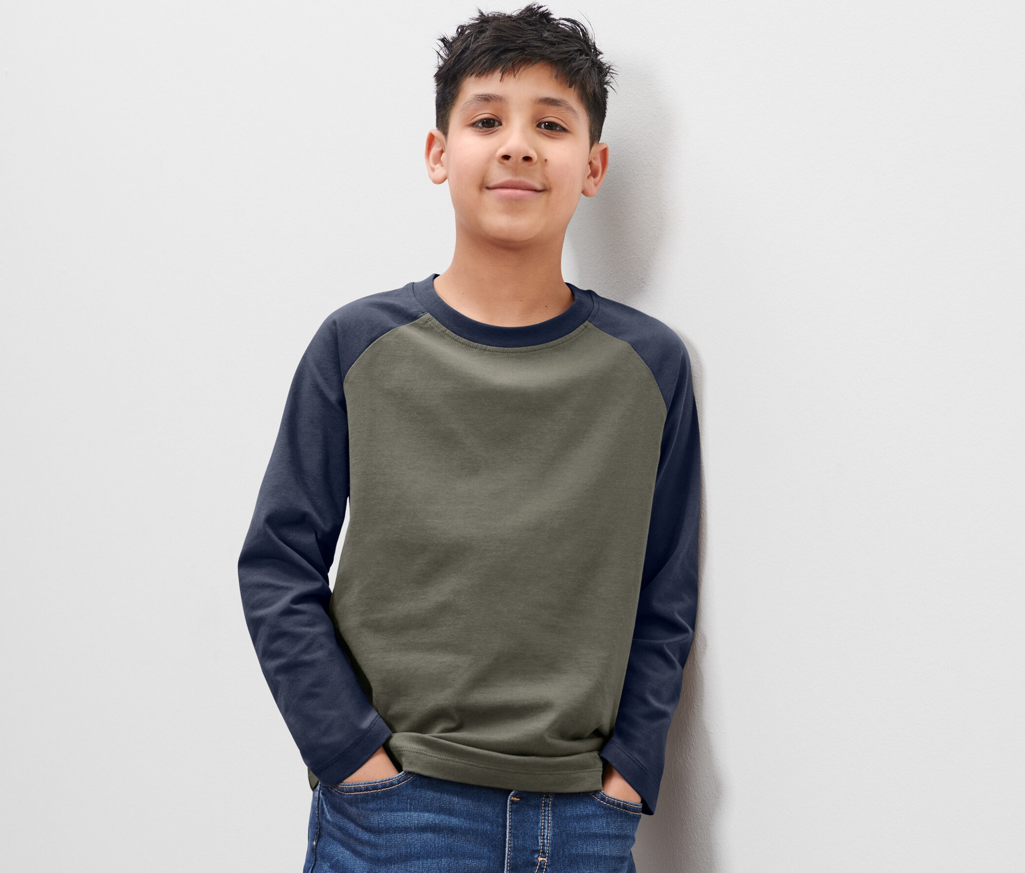 Ein Junge posiert in einem 2 Kinder-Langarmshirts und lehnt an einer weißen Wand.