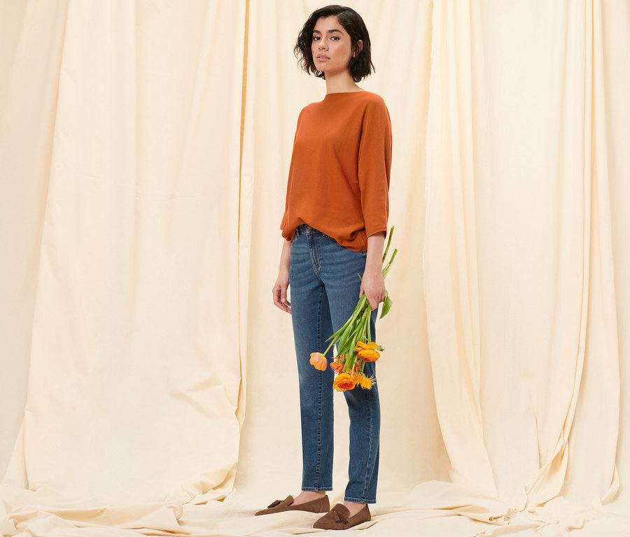 Eine Frau posiert vor einem Stoffhintergrund und hält Tulpen. Sie trägt einen orangefarbenen Feinstrickpullover, eine Slim-Denim »Fit Emma« in Midblue Denim und Loafer aus Veloursleder.