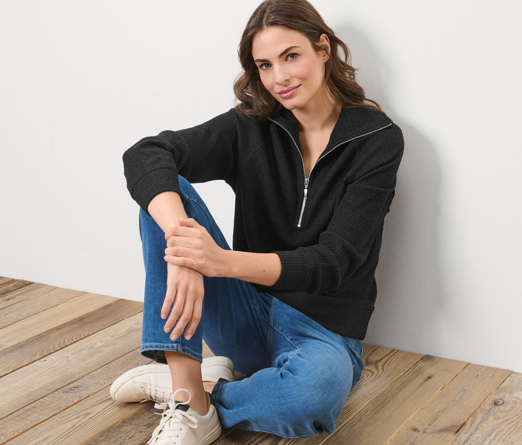 Frau sitzt auf Holzboden und trägt einen schwarzen Sweat-Troyer und Jeans.