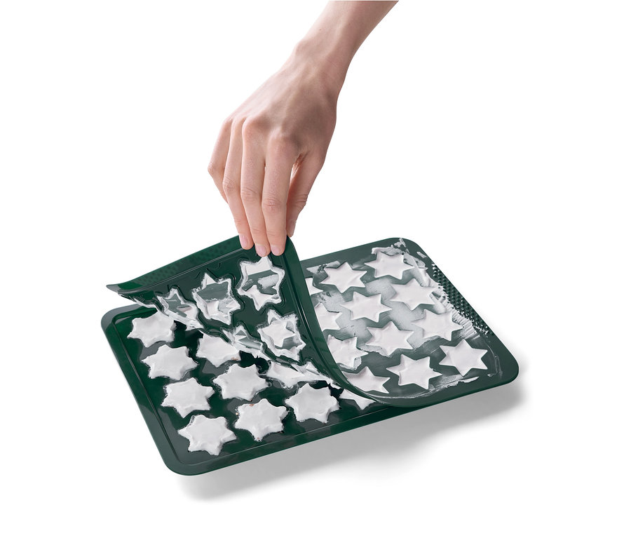 Eine Hand entfernt eine grüne Silikonform in Sternform von einem Backblech mit weißen Sternen.
