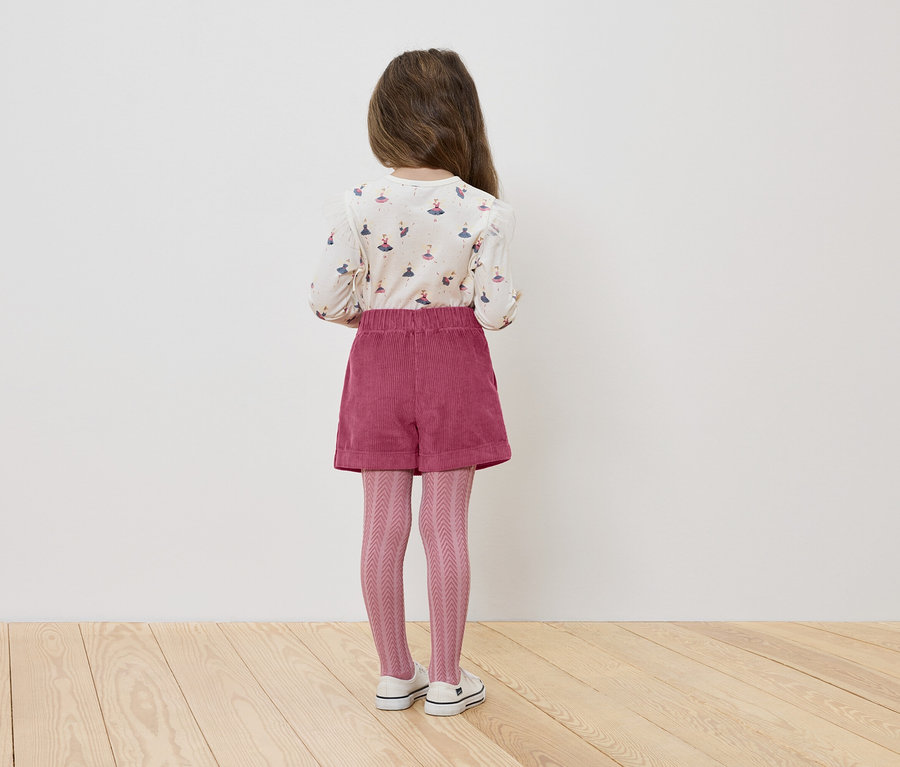 Mädchen steht mit dem Rücken zur Kamera, trägt ein Shirt mit Ballerinas, rosa Shorts und eine Strickstrumpfhose.