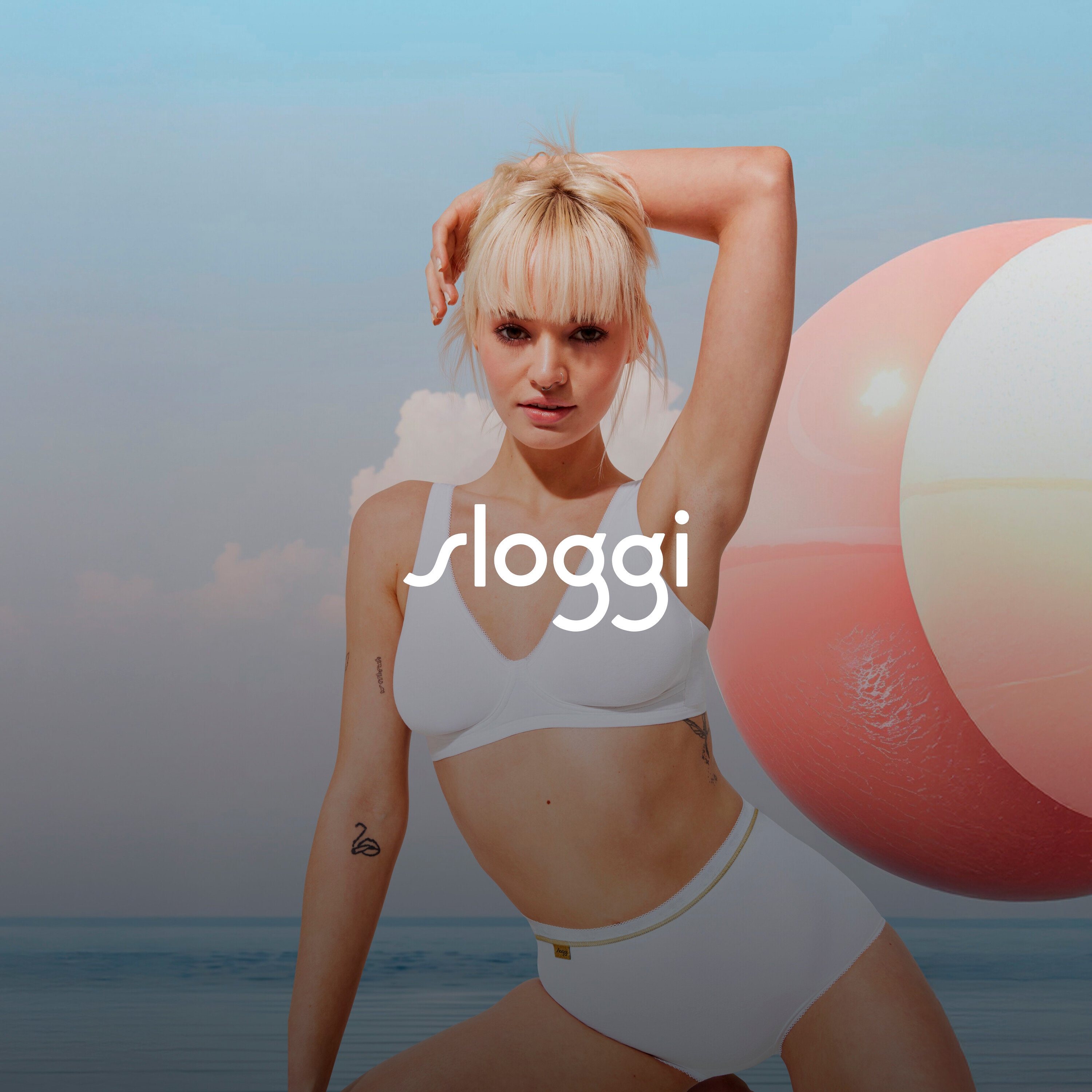 Blonde Frau in weißer Unterwäsche posiert mit einer Hand über dem Kopf neben einem Strandball.