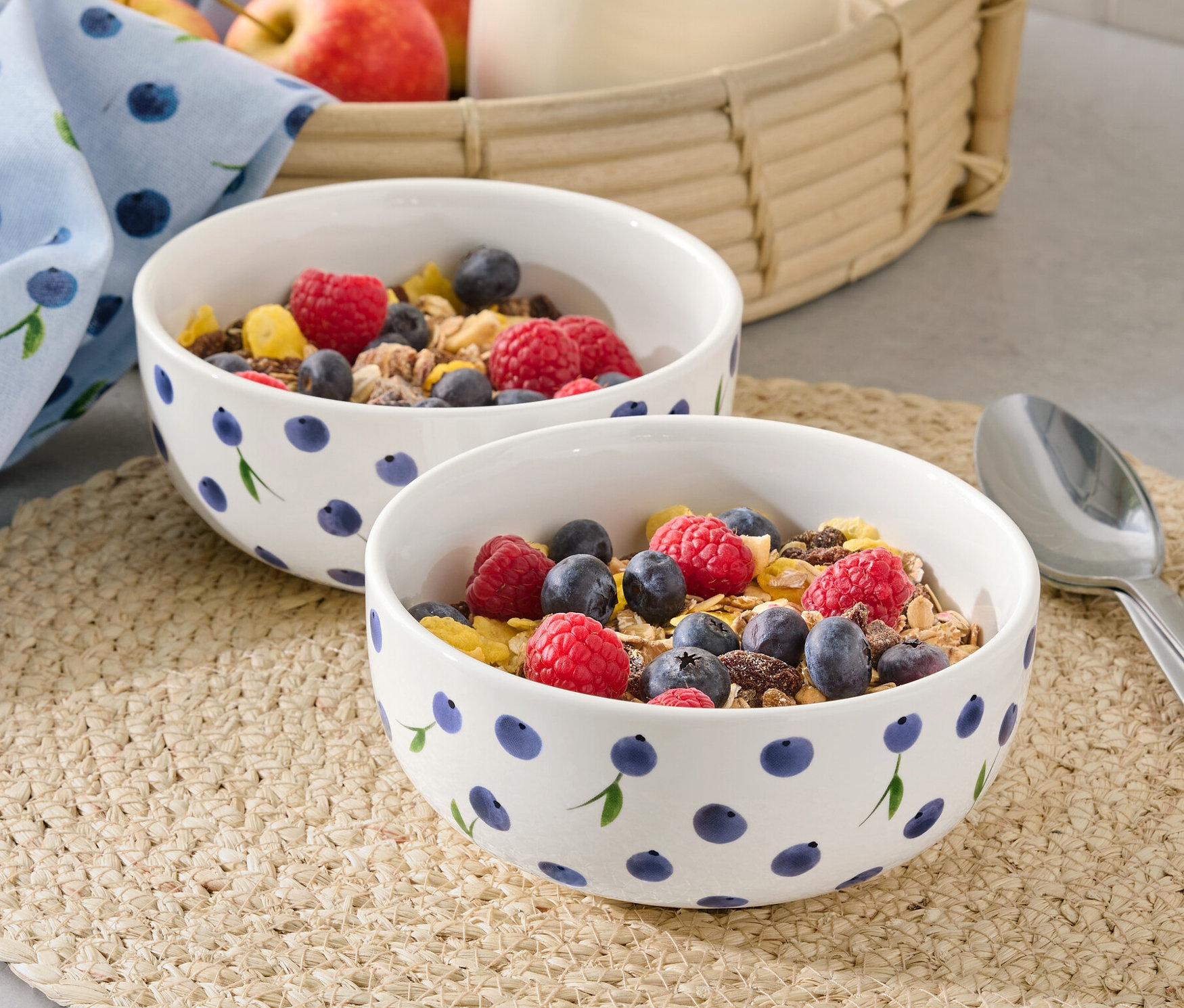 Zwei Schalen mit Müsli, Himbeeren und Blaubeeren auf einem Platzset. Im Hintergrund ein Krug mit Milch und Äpfel in einem Korb.