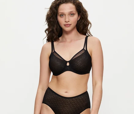 Frau mit lockigem braunem Haar trägt einen schwarzen Triumph Signature Sheer Minimizer BH und passende High-Waist-Slips, blickt in die Kamera.