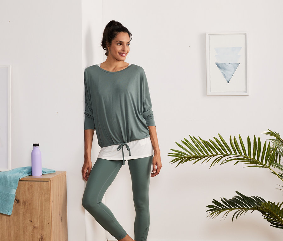 Frau lehnt an der Wand und trägt ein grün-weißes 2-in-1-Sportshirt und Leggings.