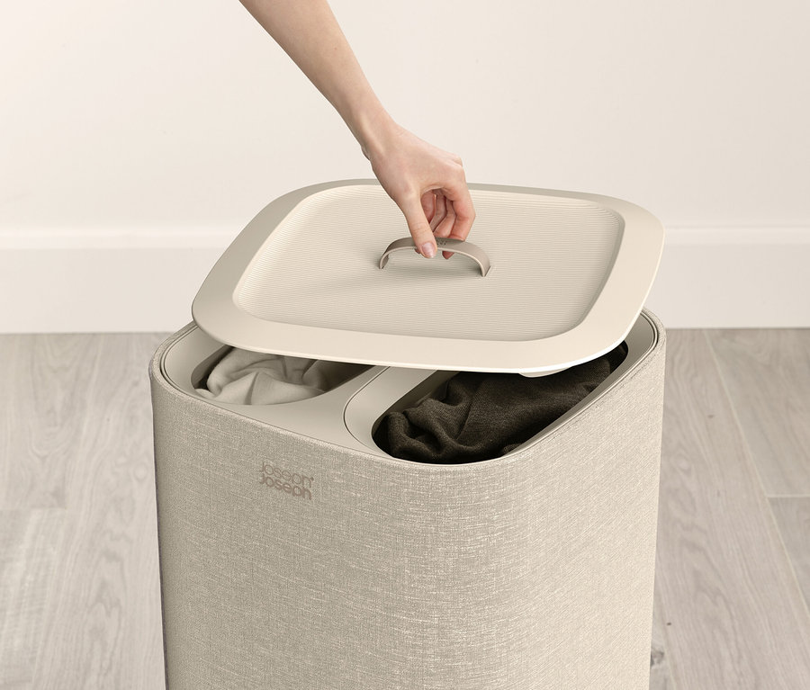 Eine Hand hebt den Deckel eines beigen Wäschekorbs mit zwei Fächern an, die jeweils mit Kleidung gefüllt sind.