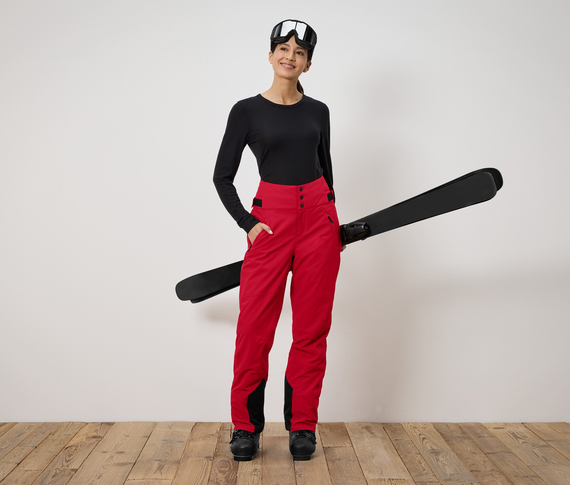 Frau steht und hält Ski. Sie trägt ein schwarzes Langarmshirt, eine rote Hose und eine Skibrille.
