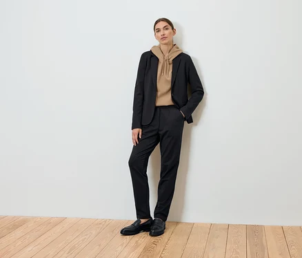 Eine Frau posiert und trägt einen schwarzen Blazer, einen braunen Kapuzenpullover und eine schwarze Hose.