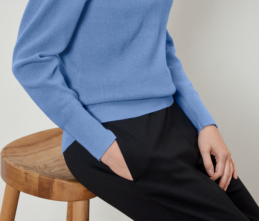 Frau sitzt auf einem Holzhocker, trägt einen blauen Pullover und eine schwarze Hose mit der Hand in der Tasche.