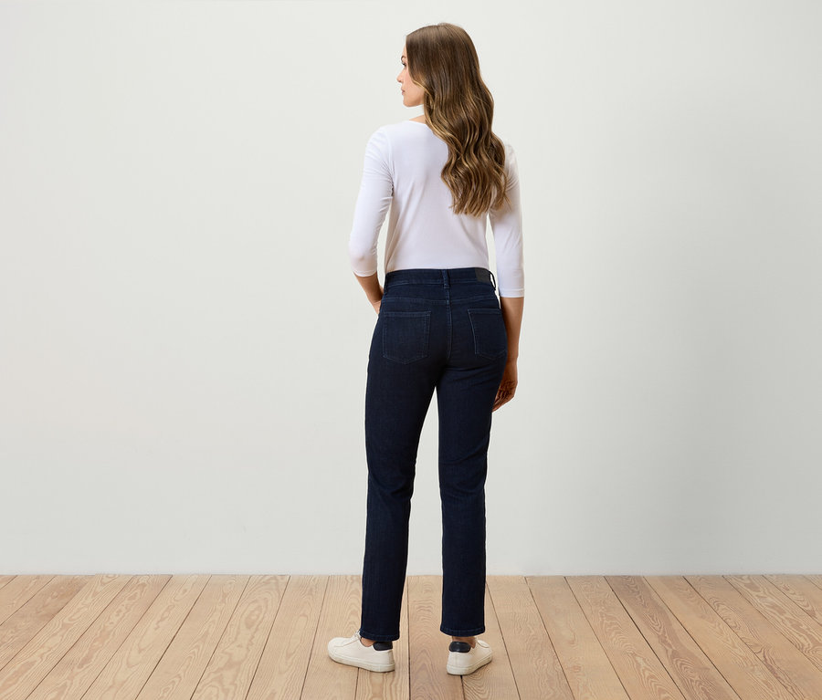 Frau steht mit dem Rücken zur Kamera und trägt dunkle Jeans, ein weißes Dreiviertelarmshirt und weiße Turnschuhe.
