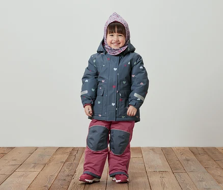 Ein lächelndes kleines Mädchen steht auf einem Holzboden und trägt eine blaue Winterjacke mit Herzmuster, eine rosafarbene Hose und ein Kopftuch.
