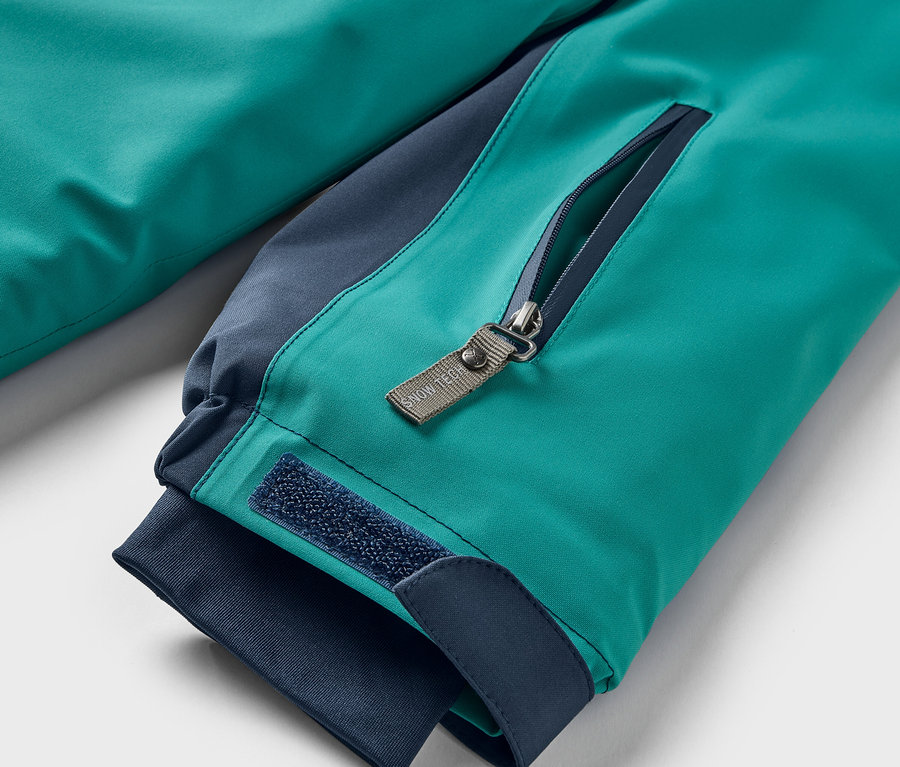Detailaufnahme einer türkisgrünen Jacke mit dunkelblauen Akzenten, Nahaufnahme einer Reißverschlusstasche und Klettverschluss am Ärmel.