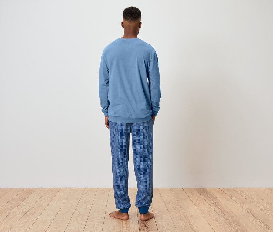 Mann von hinten trägt einen blauen Pyjama und steht auf einem Holzboden vor einer weißen Wand.