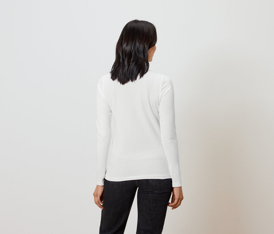 Frau mit kurzem dunklem Haar steht mit dem Rücken zur Kamera und trägt ein weißes Langarmshirt und schwarze Jeans.