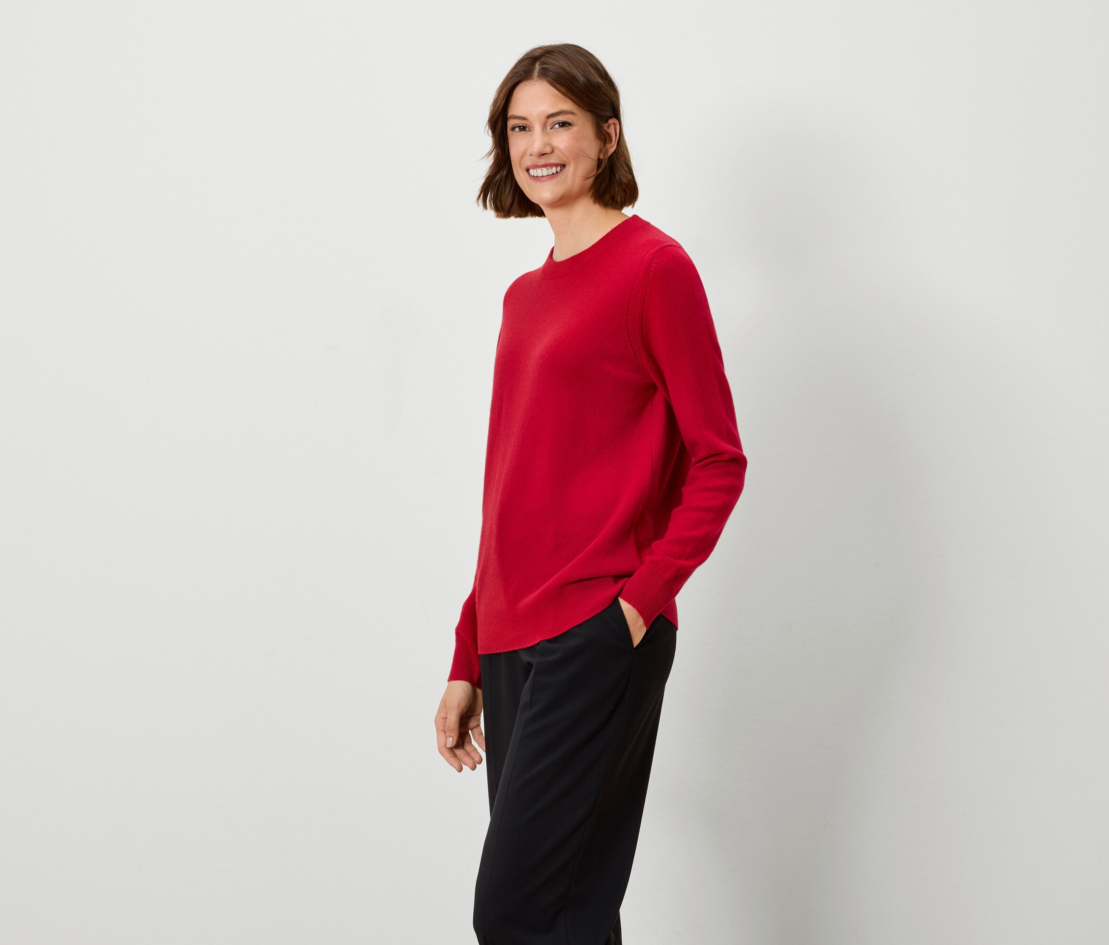 Frau mit braunen Haaren steht und trägt einen roten Pullover und eine schwarze Hose.
