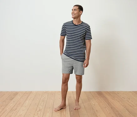 Mann steht barfuß auf einem Holzboden, trägt ein gestreiftes T-Shirt und graue Shorts.