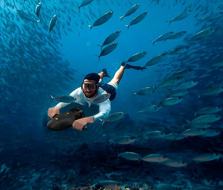 Mann schwimmt mit einem Spinera Unterwasser Powerfin und Scooter unter Wasser, umgeben von einem Fischschwarm.