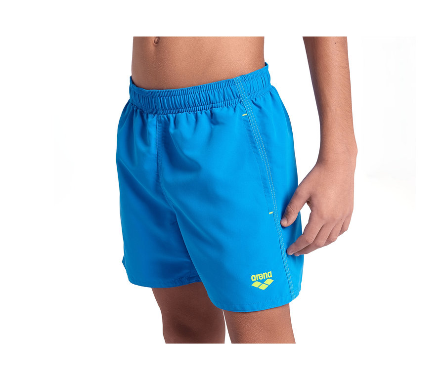 Kind trägt blaue arena Kinder-Beach Boxer Solid R Badeshorts mit gelbem arena Logo auf dem linken Bein.