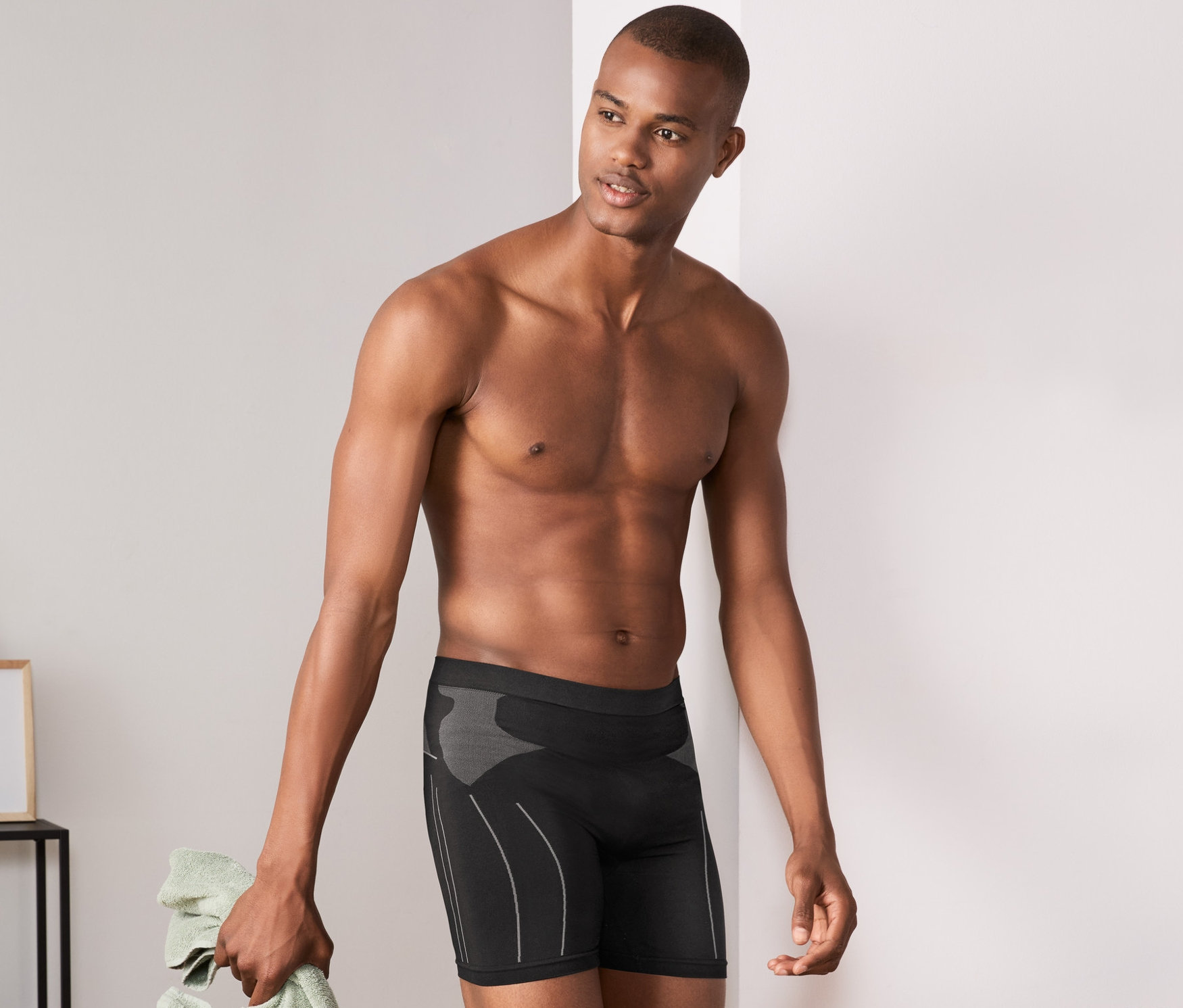 Mann lehnt an Wand, trägt Radunterhose und hält Handtuch.