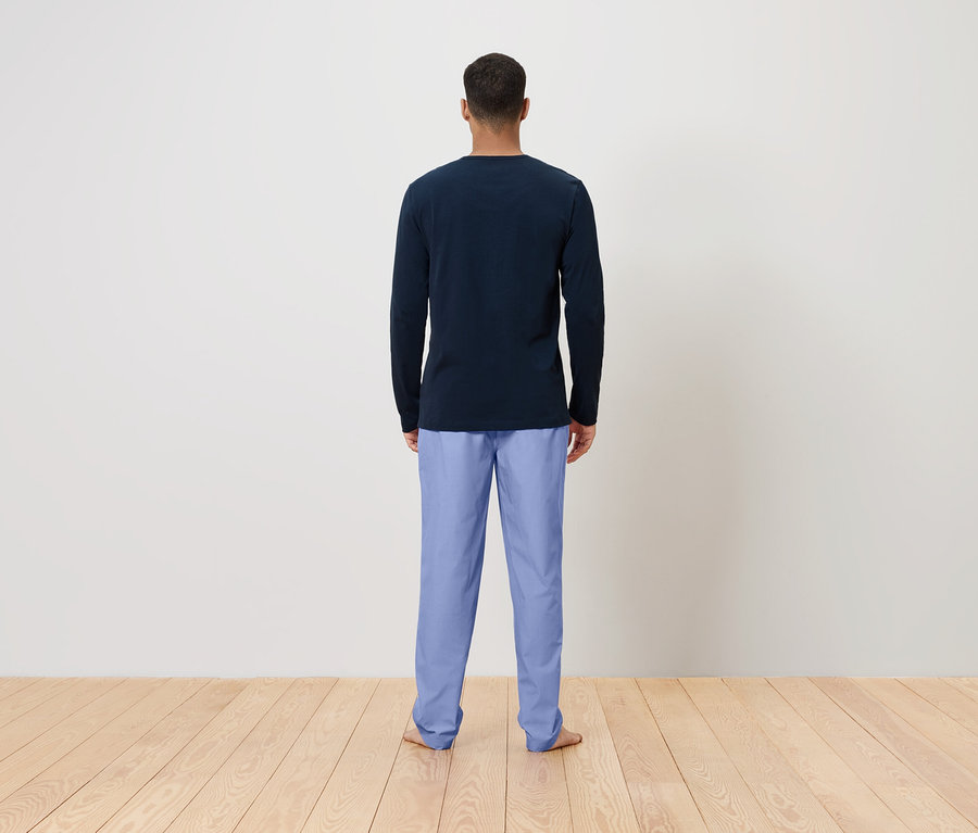 Ein Mann steht mit dem Rücken zur Kamera und trägt ein dunkelblaues langärmeliges Shirt und hellblaue Hosen.