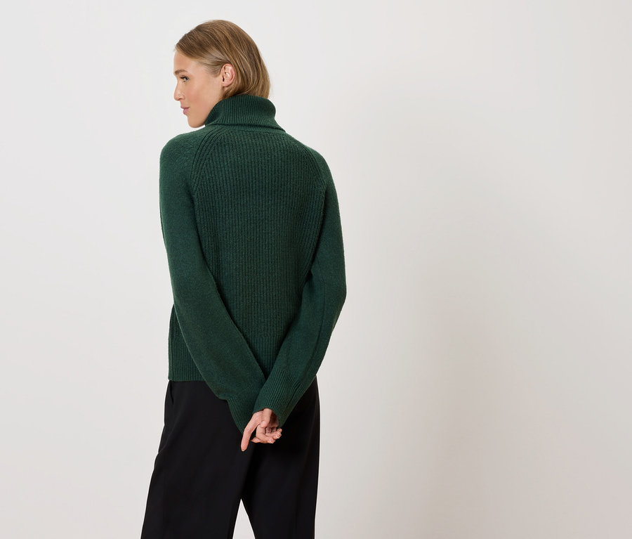 Frau mit blonden Haaren steht mit dem Rücken zur Kamera, trägt einen grünen Rollkragenpullover und eine schwarze Hose.