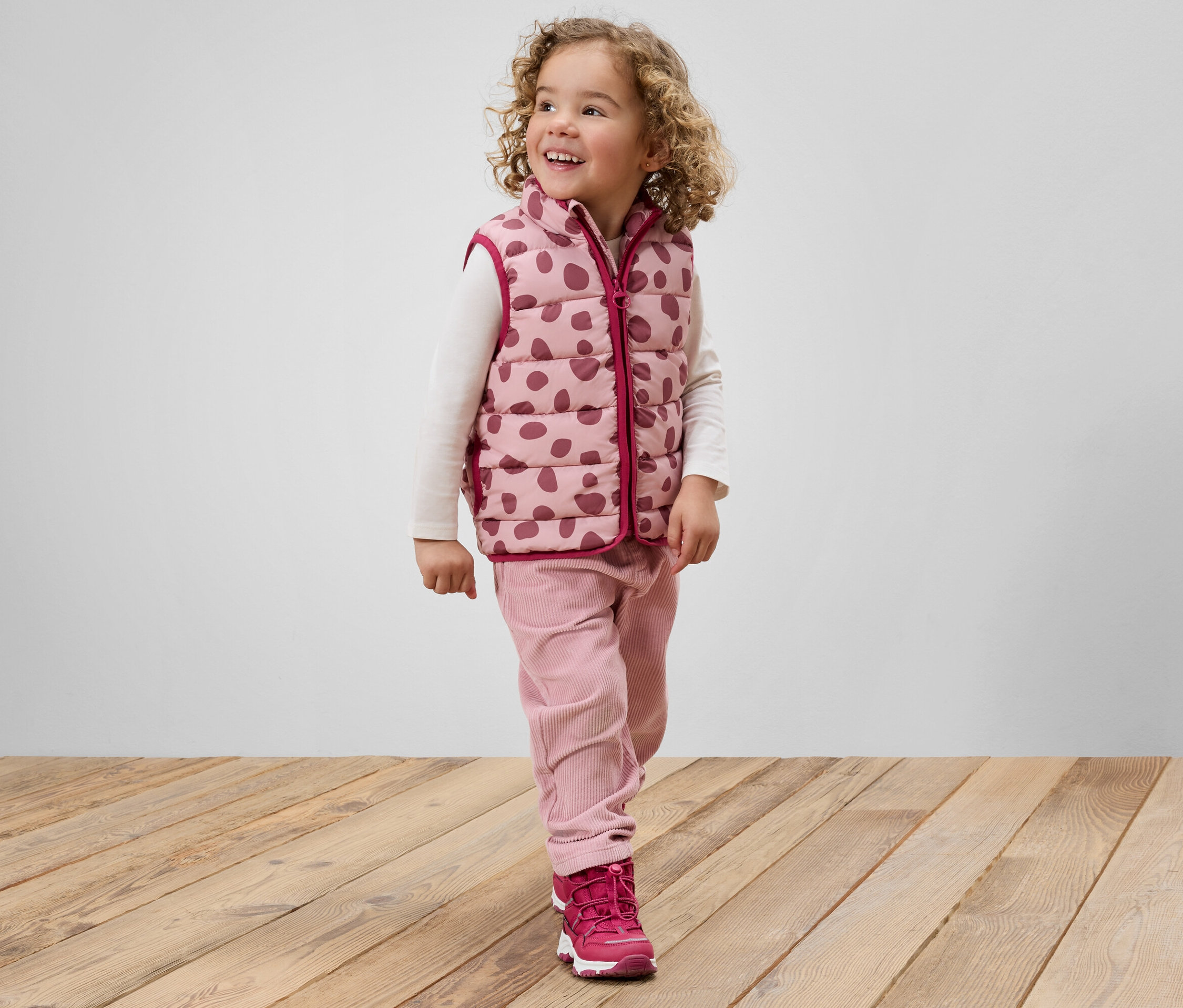 Ein lächelndes Mädchen mit lockigem Haar, gekleidet in eine rosa gepunktete Weste, rosa Hosen und rosa Schuhe, steht auf einem Holzboden.