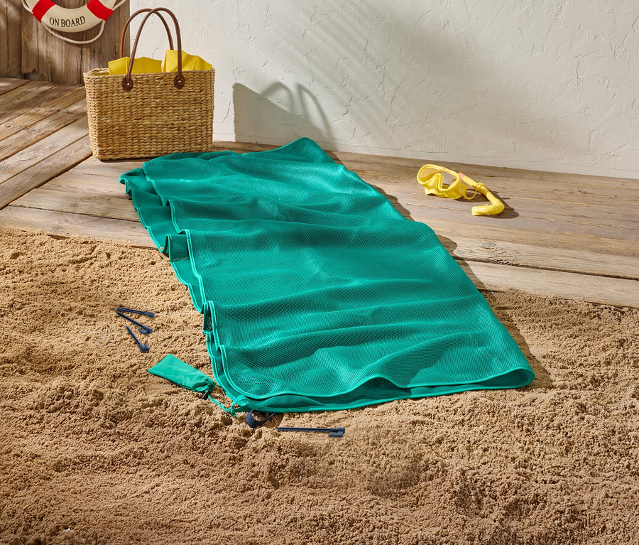 Ein grünes Strandtuch liegt auf Sand, daneben eine gelbe Taucherbrille und eine Korbtasche.