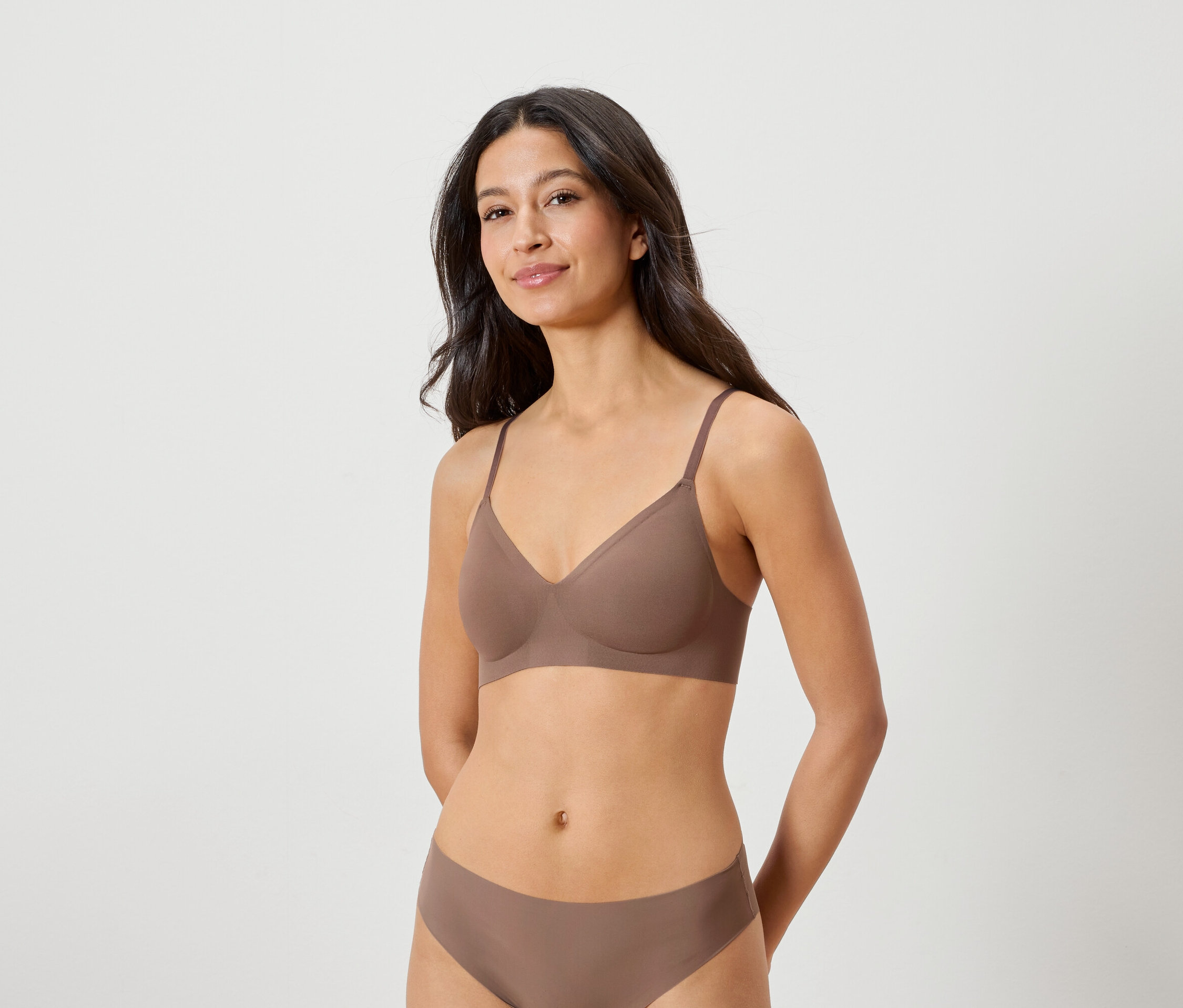 Frau mit dunklen Haaren, die einen braunen BH und Höschen trägt und mit beiden Händen hinter dem Rücken posiert.