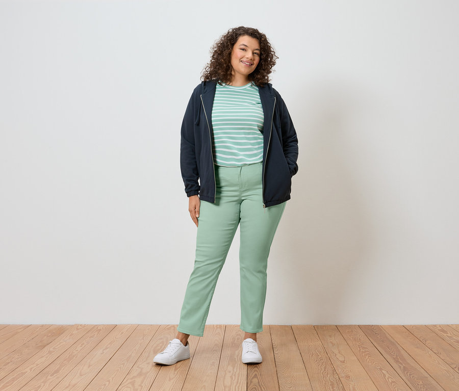 Eine Frau mit lockigem Haar steht vor einer Wand und trägt eine marineblaue Jacke, ein gestreiftes Shirt, grüne Hosen und weiße Schuhe.