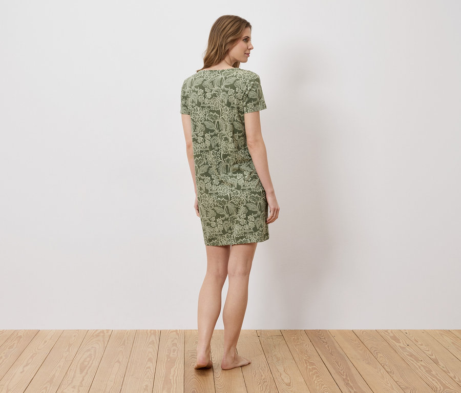 Eine Frau steht barfuß in einem grünen, kurzärmeligen Kleid mit Blumenmuster vor einer weißen Wand.