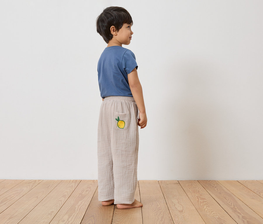 Ein Junge steht barfuß auf einem Holzboden und trägt ein blaues T-Shirt und eine lange, hellbraune Hose mit einer Zitrone auf der Tasche.