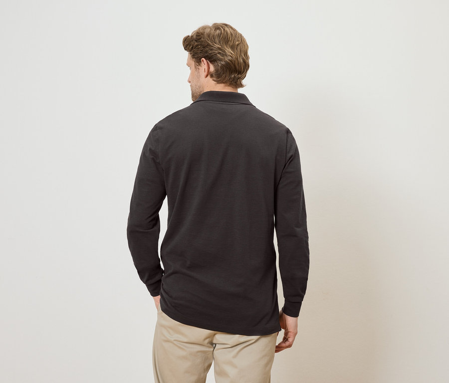 Rückansicht eines Mannes mit blonden Haaren, der ein dunkelgraues Langarm-Poloshirt und eine beige Hose vor einem cremefarbenen Hintergrund trägt.