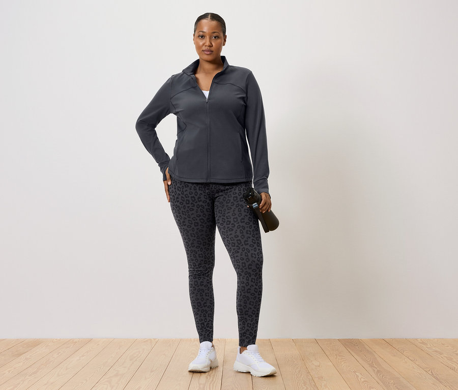 Frau in dunkler Trainingsjacke und Leggings mit Leopardenmuster, hält eine Wasserflasche.
