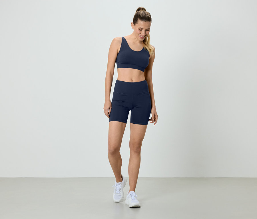 Frau in marineblauem Sport-BH und Radlerhose, mit weißen Turnschuhen, stehend vor einem weißen Hintergrund.