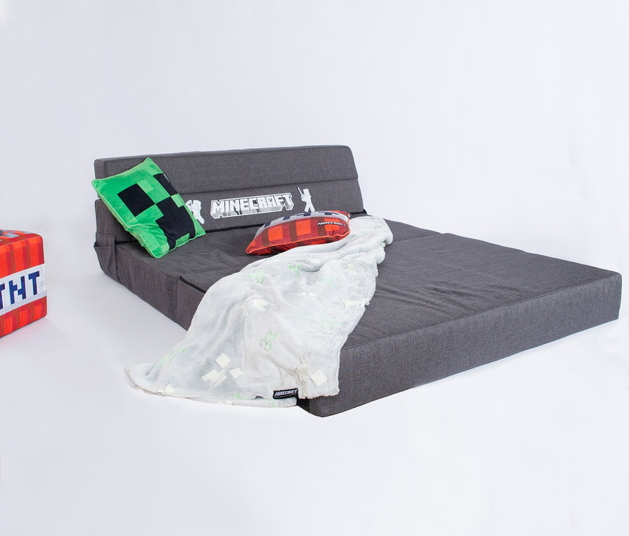 Das roba Minecraft Gaming Sofa ist ausgeklappt mit einer grauen Oberfläche. Ein grünes Minecraft-Kissen, ein rotes Minecraft-Kissen und eine weiße Decke liegen darauf.