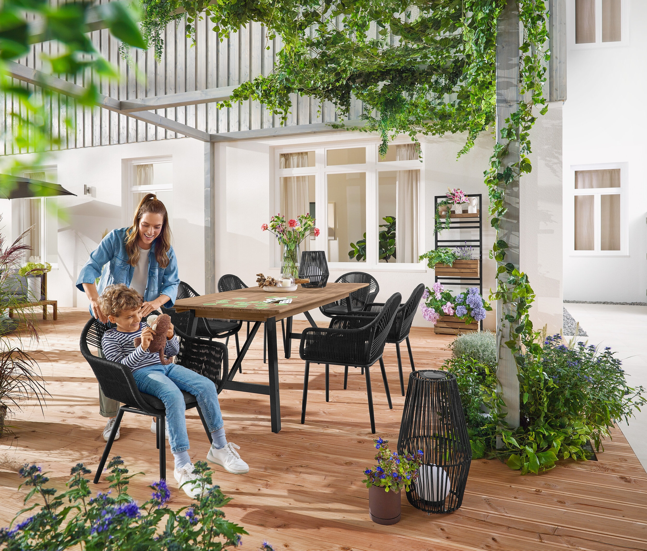 Mutter und Kind sitzen an einem Ausziehbaren Gartentisch mit rustikalem Gestell, ca. 200‒250 cm. Eine kleine und eine große Solar-Laterne mit Textilband stehen am Boden. Im Hintergrund ist eine Pflanzwand mit flexibel einhängbaren Regalen »Elin« zu sehen.