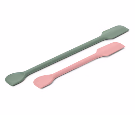 Ein grüner und ein rosa Spatel liegen diagonal auf einem weißen Hintergrund.