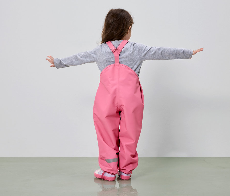 Mädchen von hinten in rosa Matschhose und grauem Oberteil mit ausgestreckten Armen vor einem hellen Hintergrund.