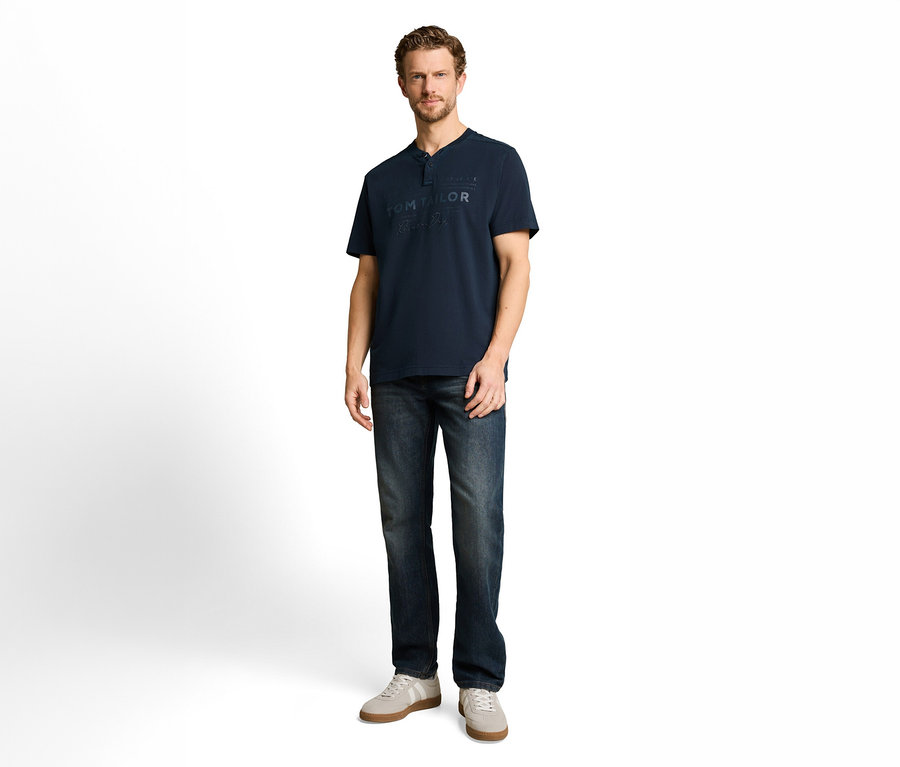 Mann in dunkelblauem Henley-Shirt mit „TOM TAILOR“-Logo, dunkler Jeans und hellgrauen Sneakers.