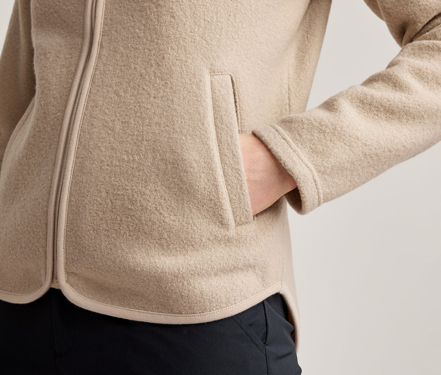Nahaufnahme einer Person in einer beigen Fleecejacke mit sichtbarem Reißverschluss und einer Hand in einer Seitentasche. Dunkle Hose unter der Jacke sichtbar.